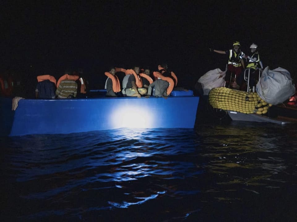 Links ein blaues Boot, worin Migranten mit Rettungswesten sitzen. Rechts ist ein Schiff, worauf Retter stehen.
