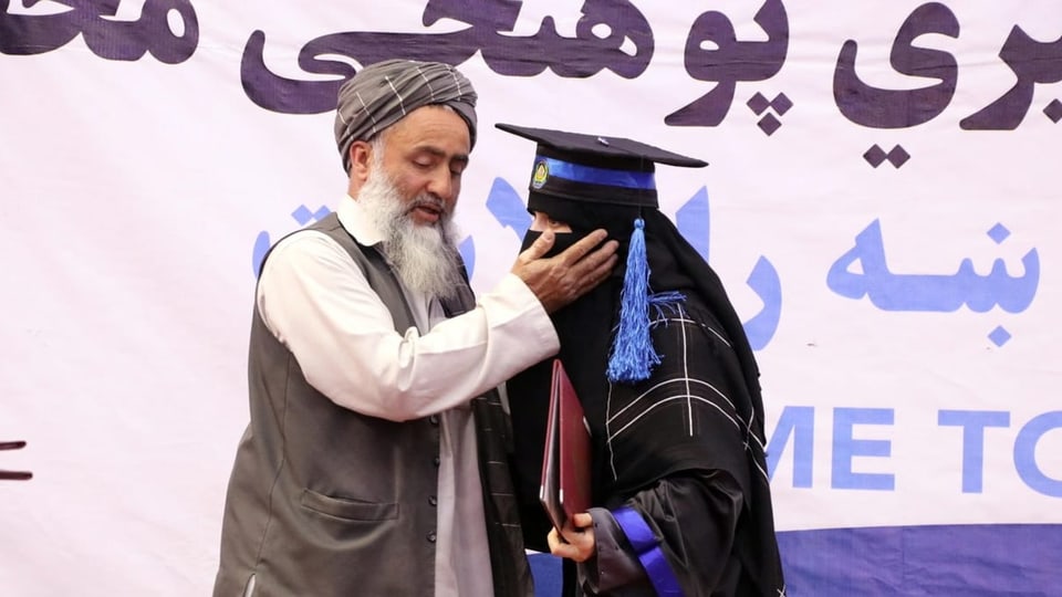 Ein Mann mit Turban berührt eine Frau mit Graduation-Hut und Verschleierung am Kinn