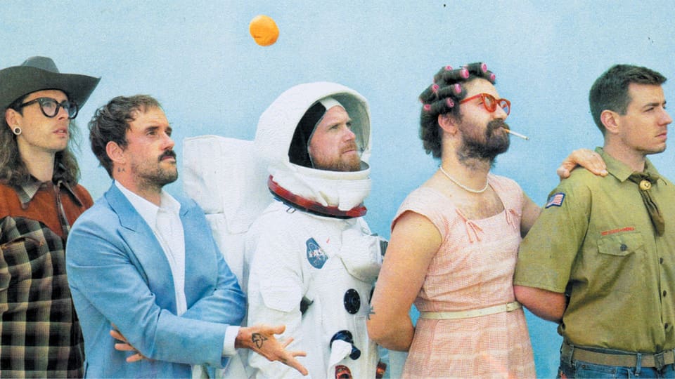 Fünf junge Männer stehen in einer Reihe und tragen spezielle Kleider: darunter Cowboyhut, Astronauten-Anzug, rosa Kleid.