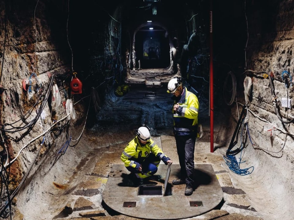 Zwei Bauarbeiter stehen in einem Tunnel auf einer Öffnung, die in ein tieferes Stockwerk führt.