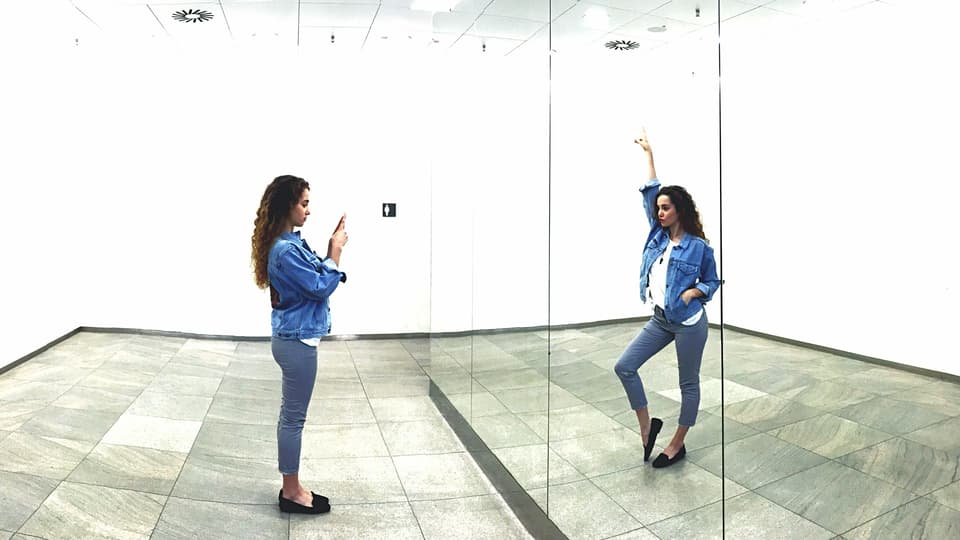 Eine Frau fotografiert sich im Spiegel. Durch eine Bildmontage macht ihr Spiegelbild eine andere Bewegung.