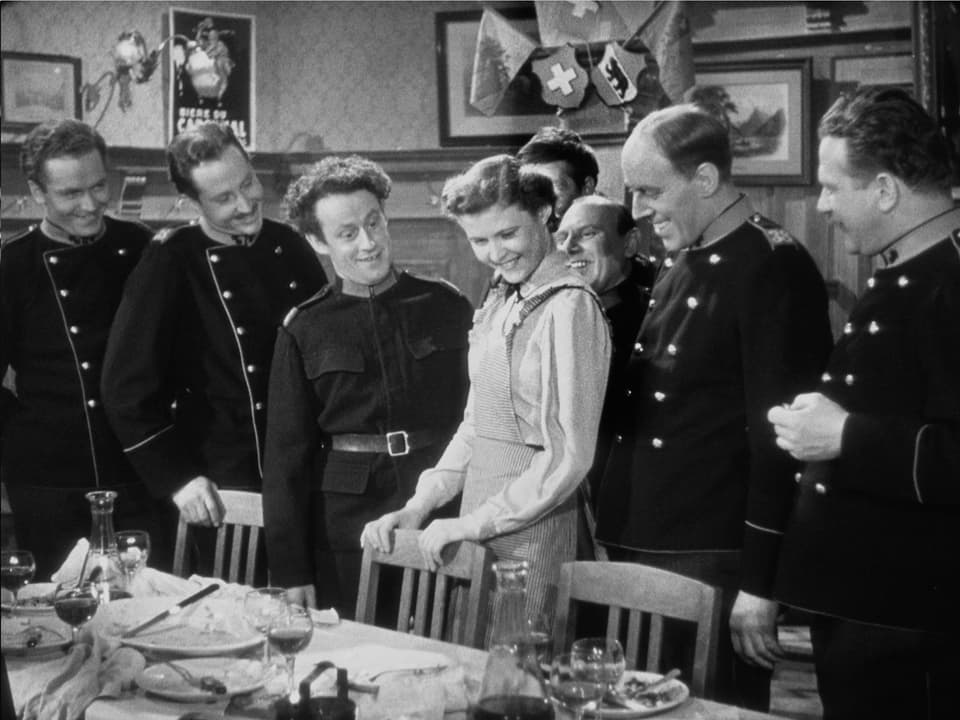 Eine Frau (Gilberte) steht an einem Tisch umgeben von sieben Soldaten