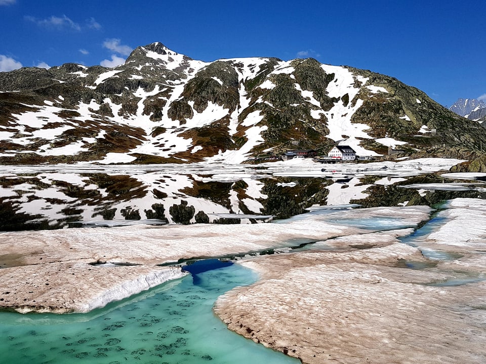 4. Juli Im Vordergrund ein Bergsee mit Schnee und Eis, im Hintergrund Berge und blauer Himmel.