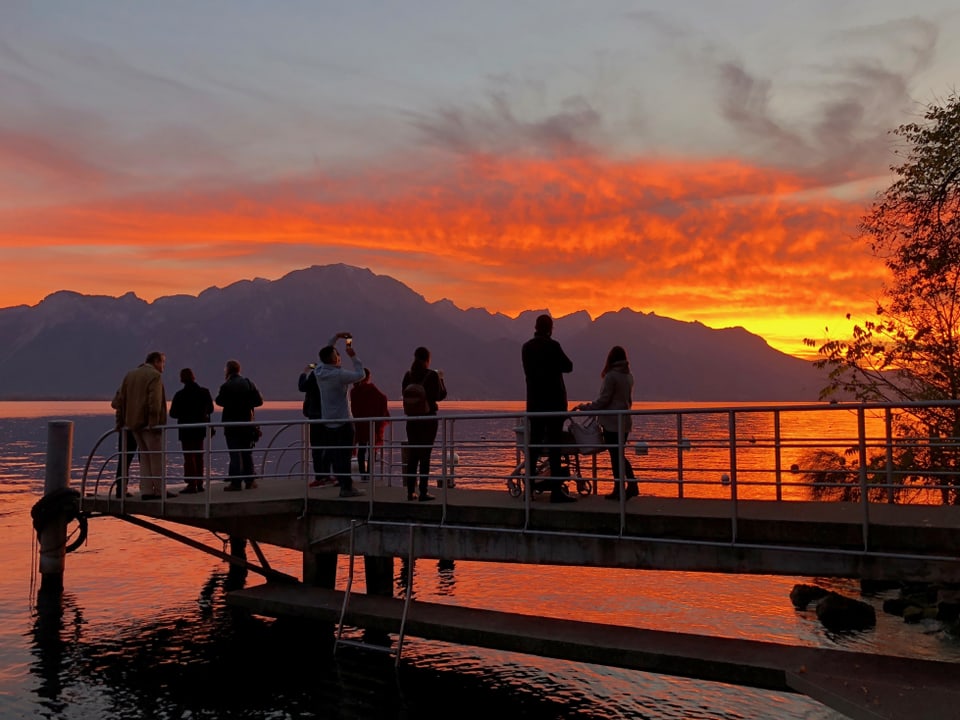 Personengruppe auf dem Schiffssteg bei Montreux, dahinter der rote See und der rote Himmel.