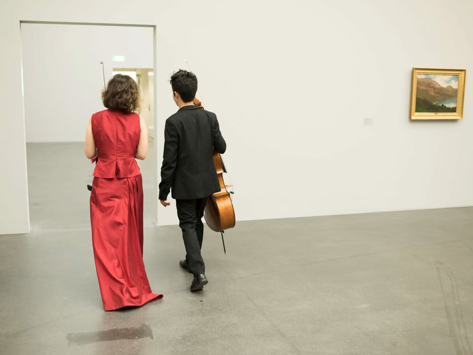 Eine Frau und ein Mann von hinten in einem Museumsraum. An der rechten Wand hängt ein Landschaftsgemälde mit Goldrahmen. Sie trägt ein rotes Abendkleid, er einen schwarzen Anzug. Beide haben ihre Instrumente in den Händen. Sie laufen nebeneinander von einem Museumsraum in den nächsten. Sie sind von hinten abgebildet.