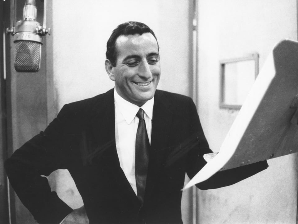 schwarz-weiss Bild mit einem Mann mit dunklen Haaren der neben einem Notenständer und einem Mikrofon steht und lächelt.