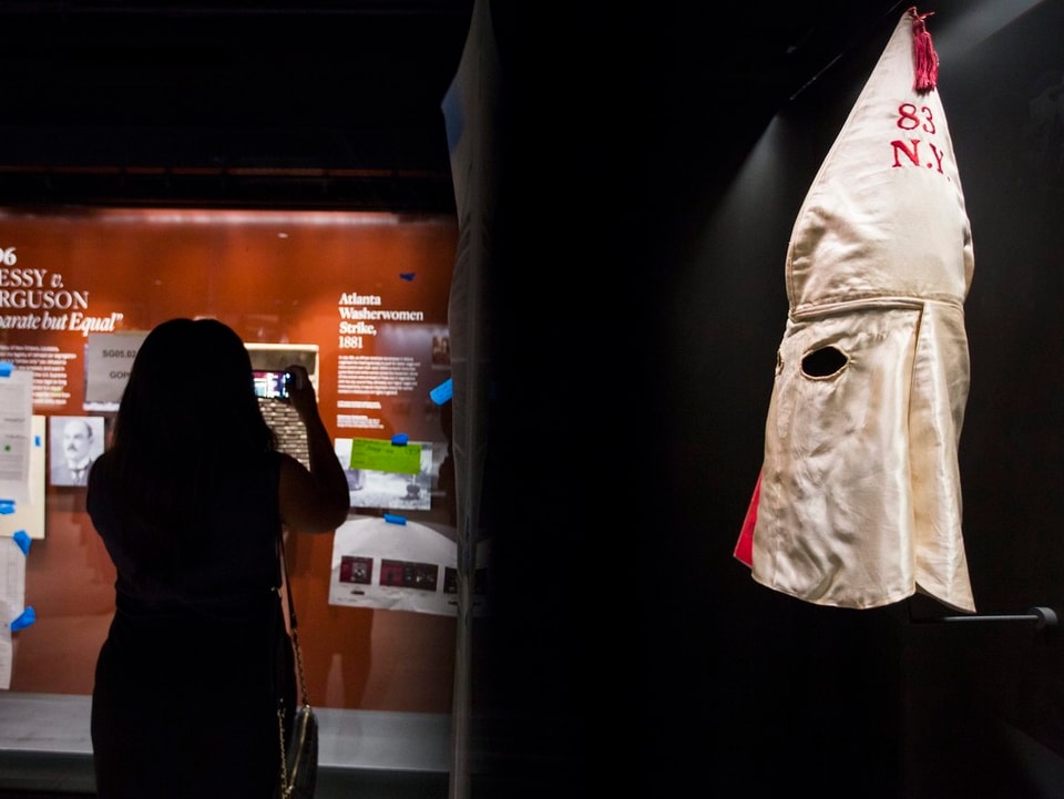 Ku-Klux-Klan-Maske hängt an einer Wand, dahinter fotografierende Frau im Museum.