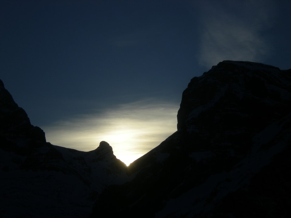 Der Himmel ist mehrheitlich blau. Unten sieht man die Silouhette eines Bergmassivs. Die Sonne ist knapp hinter einer Bergkante zu erkennen.