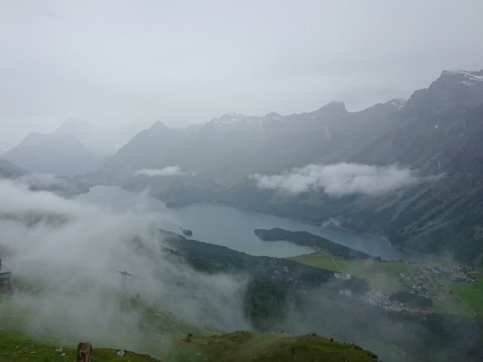 Wolkenverhangenes Bild in grautönen. Knapp zu erkennen ist ein Alpental mit grünen Wiesen und einem See. Dahinter schroffe Bergflanken, verschwommen im Nebel. 