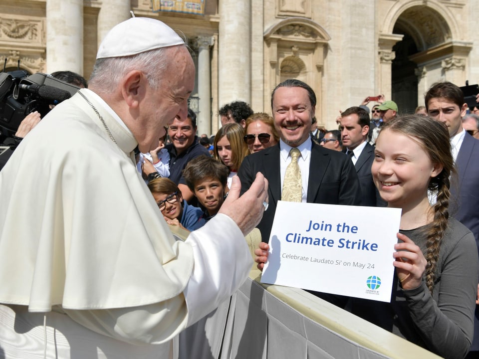 Papst Franziskus spricht mit und Greta Thunberg, die hinter einer Absperrung auf dem Petersplatz steht; sie hält ein Blatt Papier mit der Aufschrift «Join the Climate Strike» hoch. Beide lächeln.