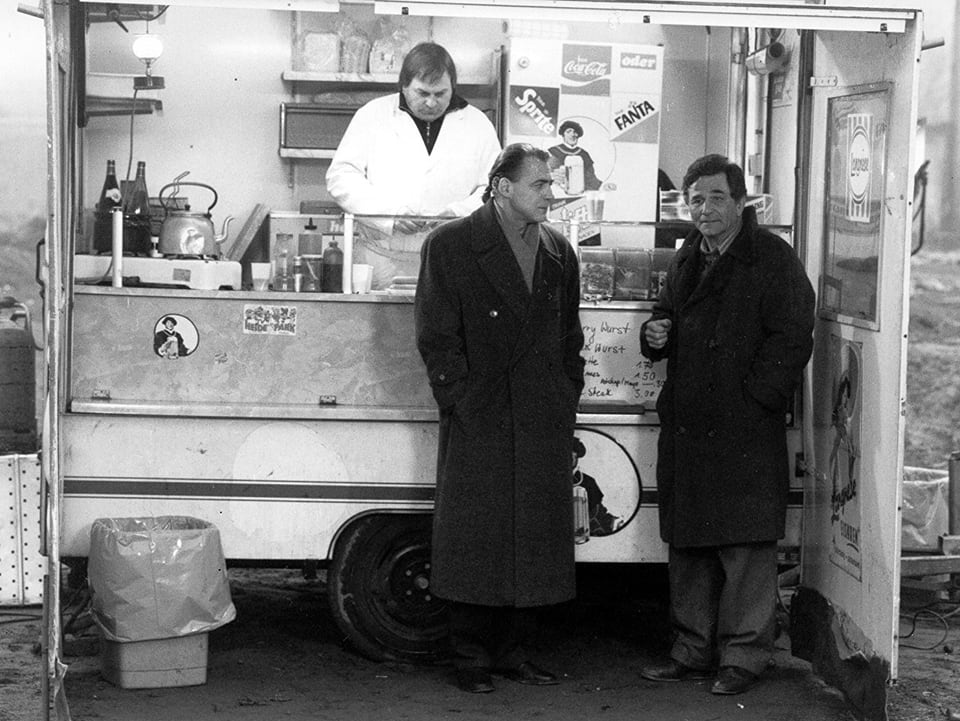 Schwarzweissbild: Zwei Männer in Mänteln stehen an einem Imbissstand