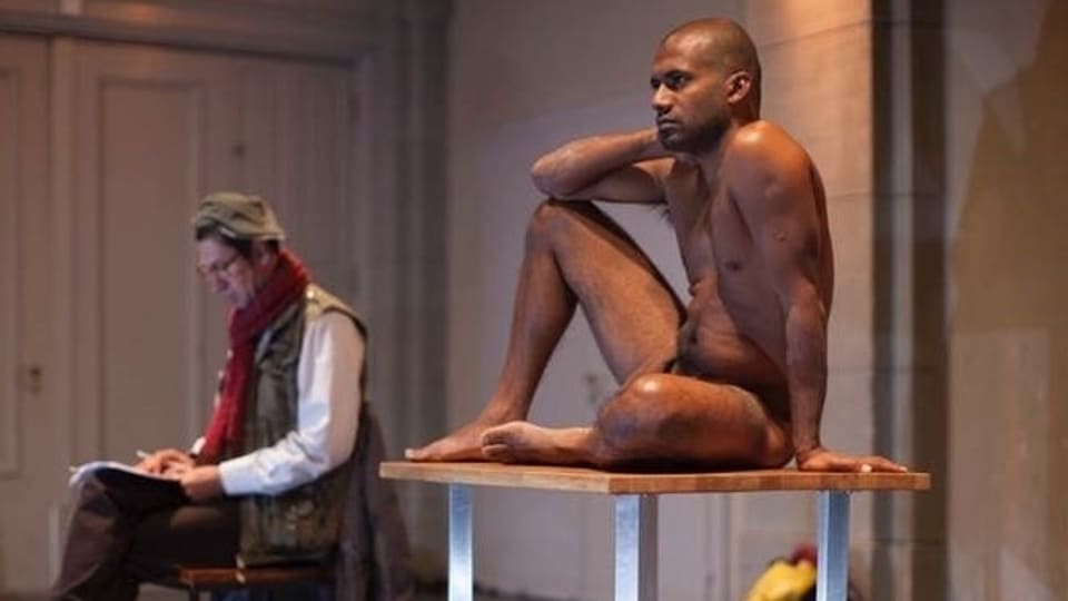 Theaterszene: Ein Mann mit rotem Schal sitzt auf einem Stuhl, ein dunkelhäutiger Mann nackt auf einem Tisch.