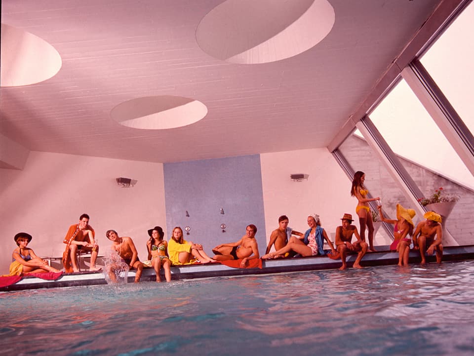 Ein Indoor-Swimmingpool mit futuristischer Decke. Viele Menschen sitzen am Rand.