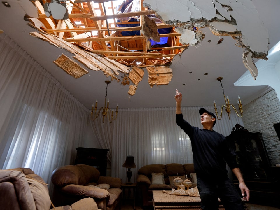 Ein Bewohner steht in seinem Wohnzimmer und zeigt auf die Decke. Diese ist zerstört, man sieht ins obere Stockwerk.