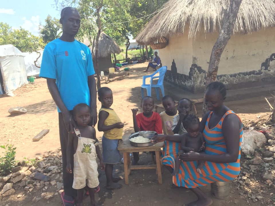 Ein Südsudanese steht neben seiner Familie, die Hände auf den Schultern eines Kleinkindes. Daneben sitzen seine Frau und die fünf anderen Kinder rund um einen Tisch und essen aus derselben Schüssel.