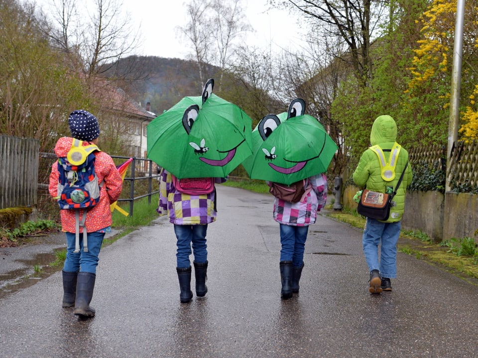 Vier Kinder laufen mit grünen Froschschirmen und farbigen Jacken in die Schule.
