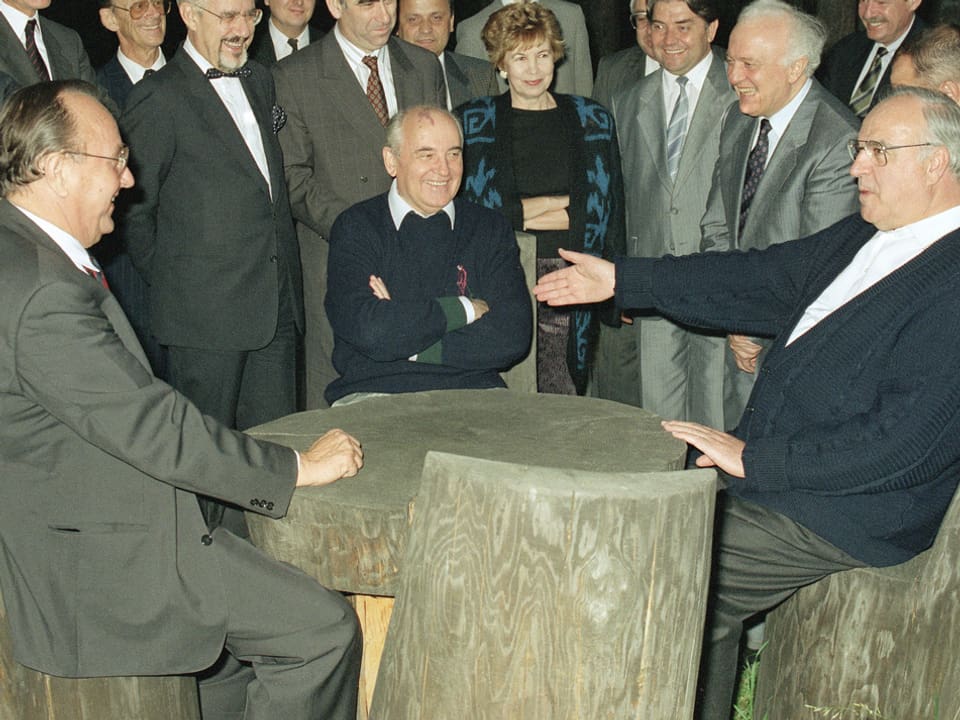 Genscher und der sowjetischen Präsident Michail Gorbatschow (Mitte) und der deutsche Kanzler Helmut Kohl sitzen während des historischen Treffens beider Länder im Kaukasus an einem Holztisch auf Stühlen aus Baumstämmen