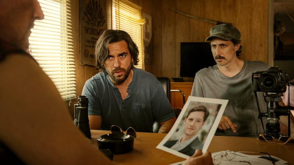 Ein Mann mit längerem, dunklem Haar und einer mit Kappe schauen auf einen Mann am Tisch, hält ein Foto.