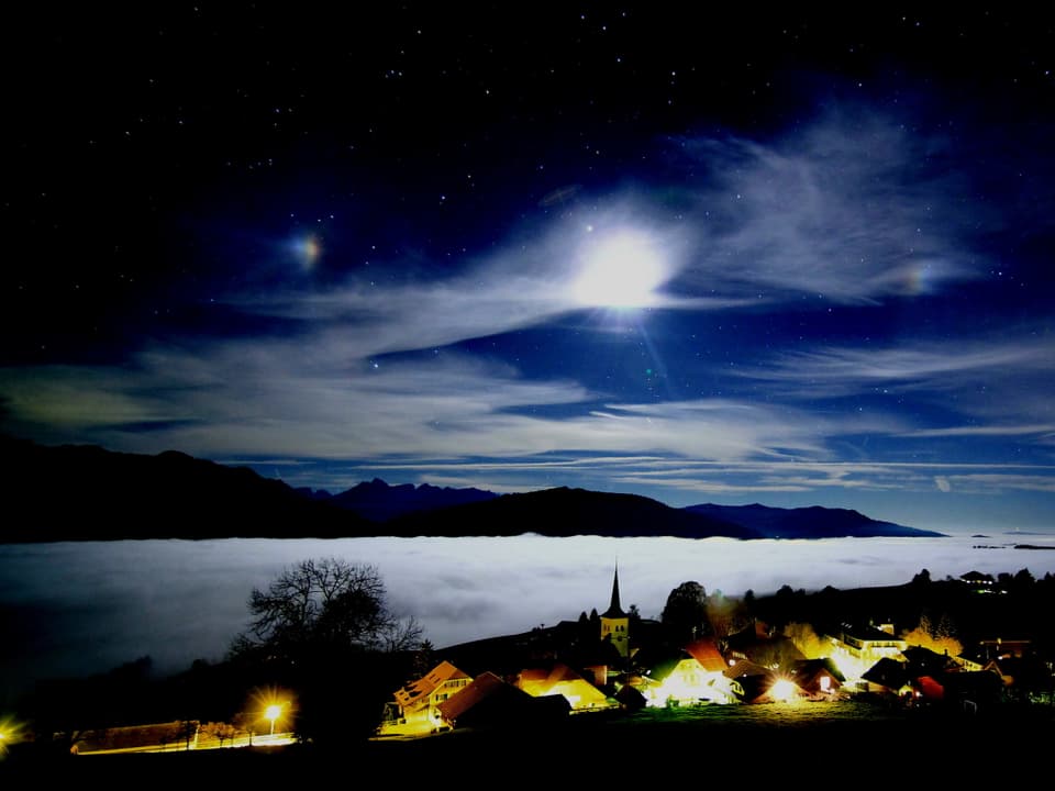 Nacht mit leuchtendem Dorf. Dahinter Nebel im Tal. Am Himmel wenige Wolken und Mond. In den Wolken Haloeffekt. 