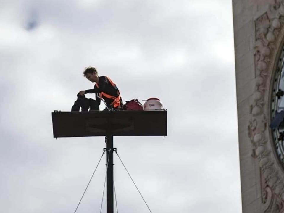 Ein Mann sitzt auf einer rechteckigen Platte, die 20 Meter über dem Boden an einem Masten festgemacht ist.