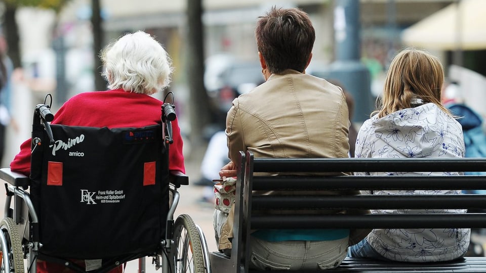 Eine alte Frau in einem Rollstuhl, eine älter Frau und ein Mädchen auf einer Parkbank nebeneinander.