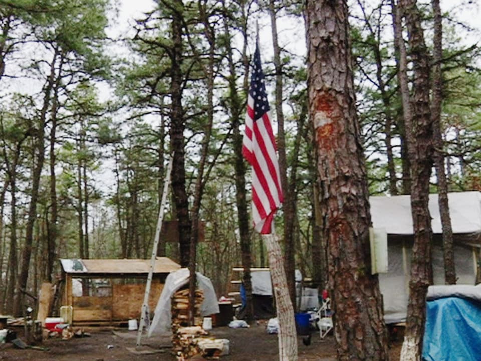 Improvisierte Baracke in einem Wald. An einem Baum hängt eine US-amerikanische Flagge