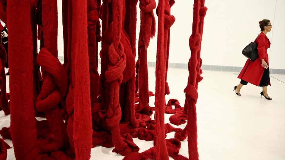 Rote Wollfäden hängen von Decke, Frau in rotem Mantel geht daran vrobei.