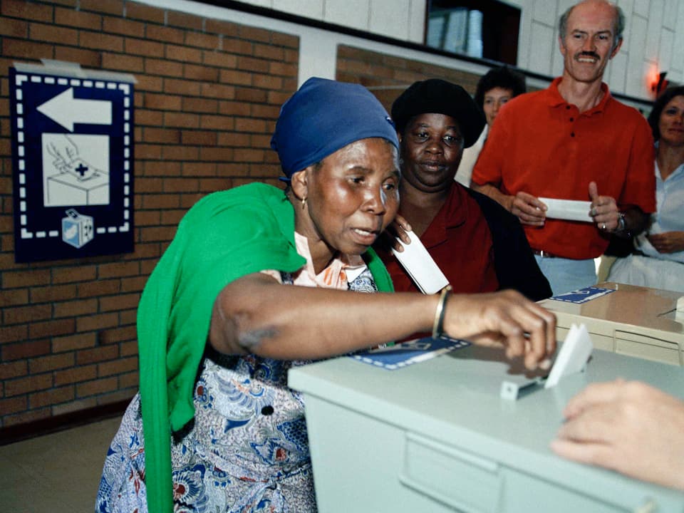 Eine ältere schwarze Frau schiebt unsicher einen Wahlzettel in eine Urne, auch ein Weisser steht an.