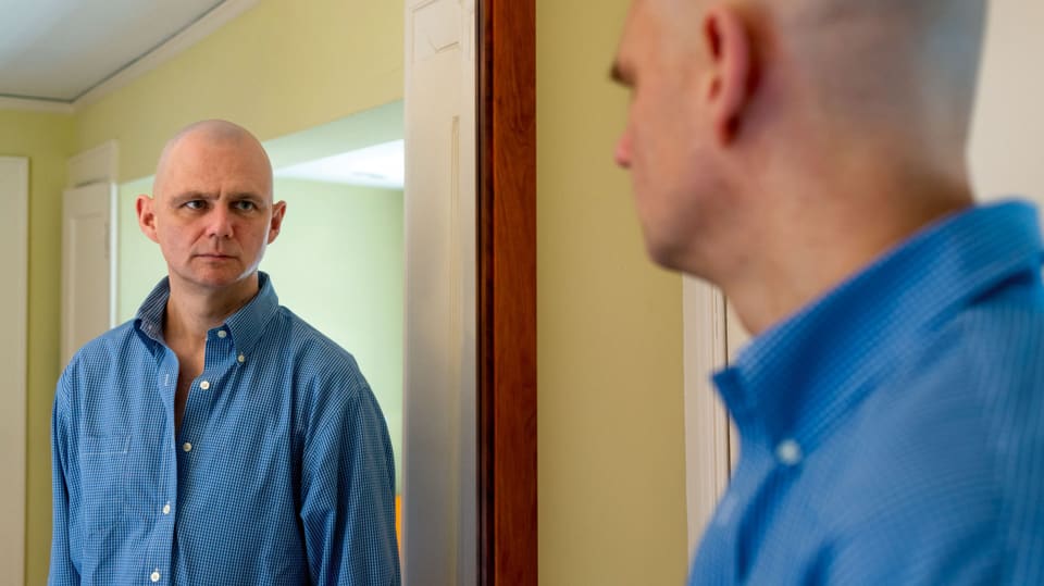 Ein Mann in blauem Hemd und ohne Haare blickt in einen Spiegel.