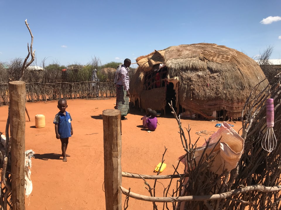 Kleine Kinder spielen vor einer runden Hütte, eine Frau schaut zu.