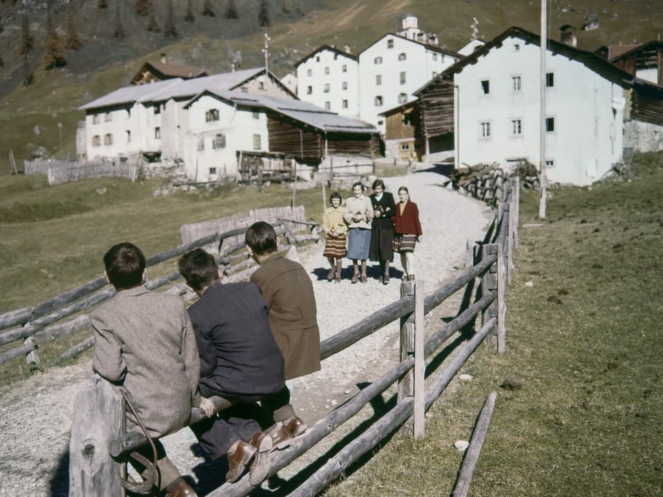 Bild aus den 50er Jahren. Drei Männer beobachten vier vorbeilaufende Frauen in einem Bergdorf.