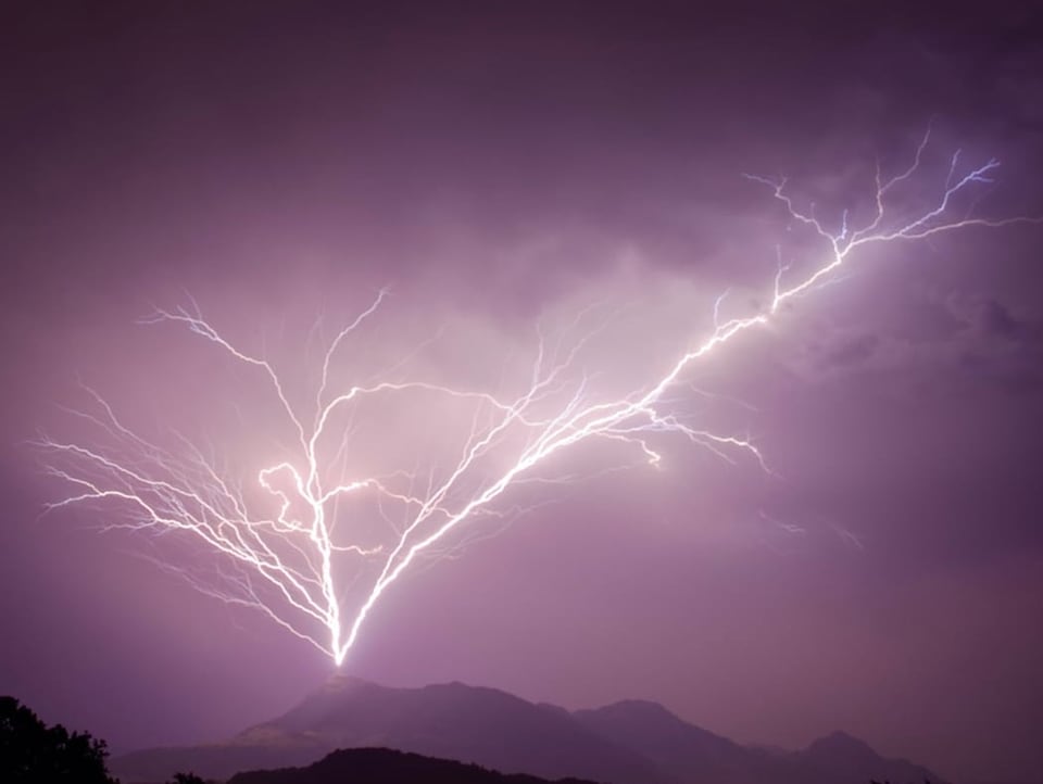 Dunkles Foto in violett, das einen Berggipfel zeigt, vom dem aus einige Blitze in den Himmel hinauf ragen. 