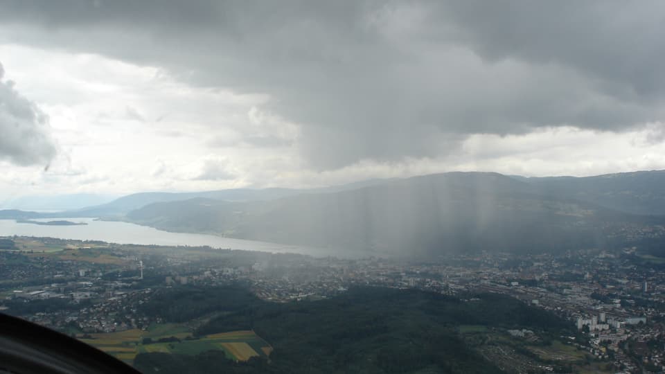 Flugbild: Landschaft mit See und Jurahügeln im Hintergrund. Im Vordergrund graue Wolke mit fallenden Regenstreifen. 