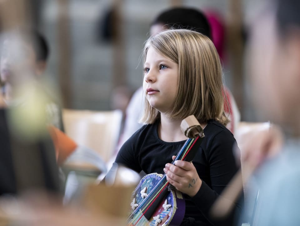 Ein kleines Mädchen hält eine selbstgebastelte Geige in der Hand und hört dem Lehrer zu.