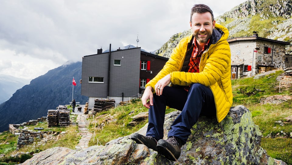 Nik Hartmann sitzt auf einem Stein, im Hintergrund ist eine Hütte zu sehen.