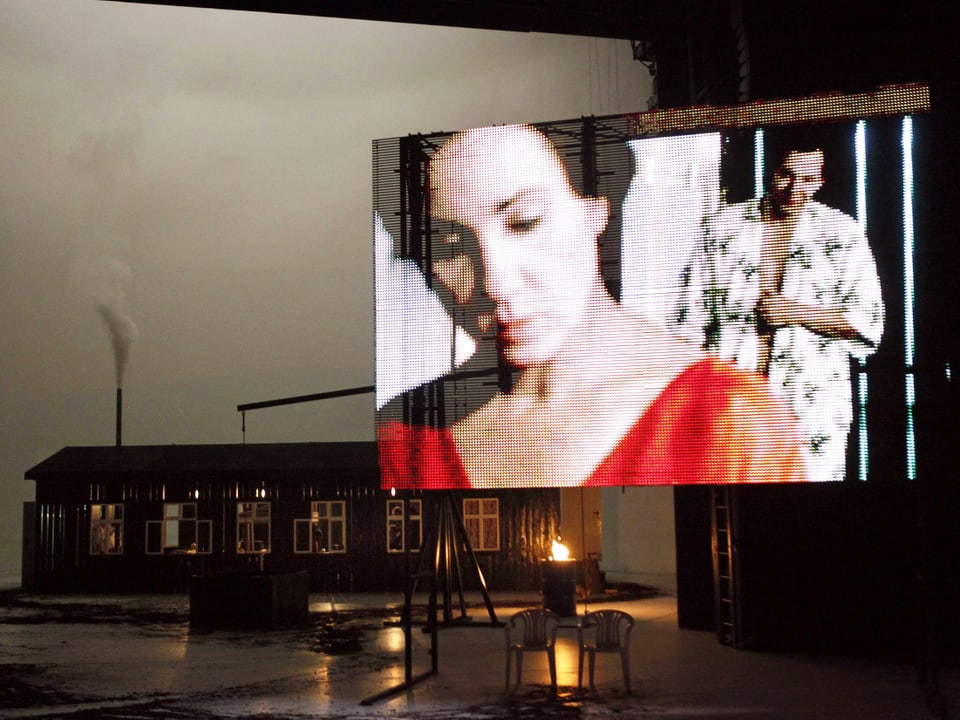 Eine Frau und ein Mann auf einer Leinwand auf einer kargen Theaterbühne.
