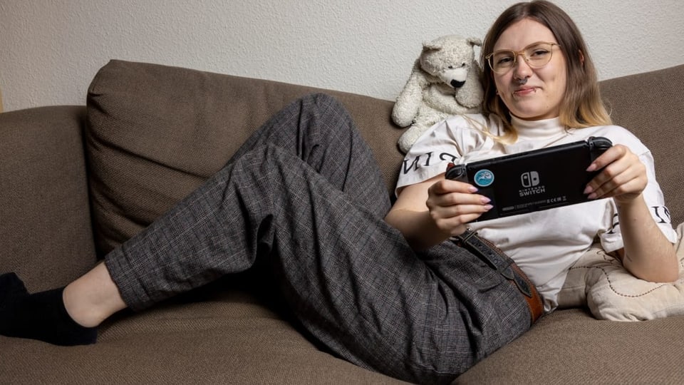 junge Frau mit Lippenpiercing, Nasenring, heller Brille auf der Couch, hält Gamekonsole, Beine auf dem Sofa.