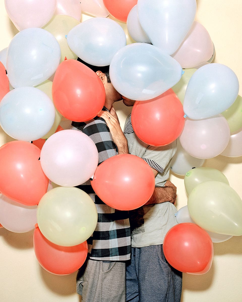 Zwei Männer küssen sich und sind dabei hinter Luftballonen versteckt.