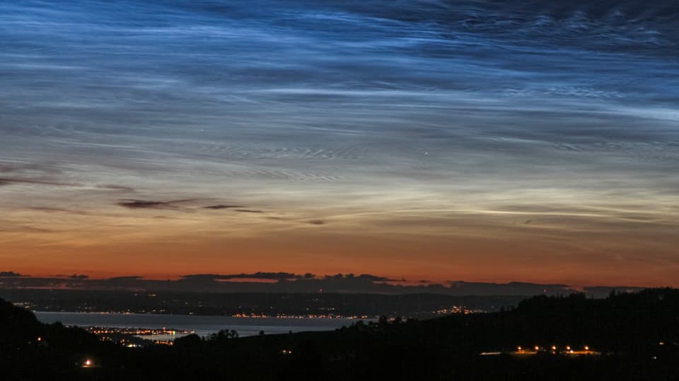 Leuchtende Nachtwolken Blick zum Bodensee, darüber das Abendrot mit dunklen Cirren, darüber die bläulich-weissen Leuchtenden Nachtwolken.