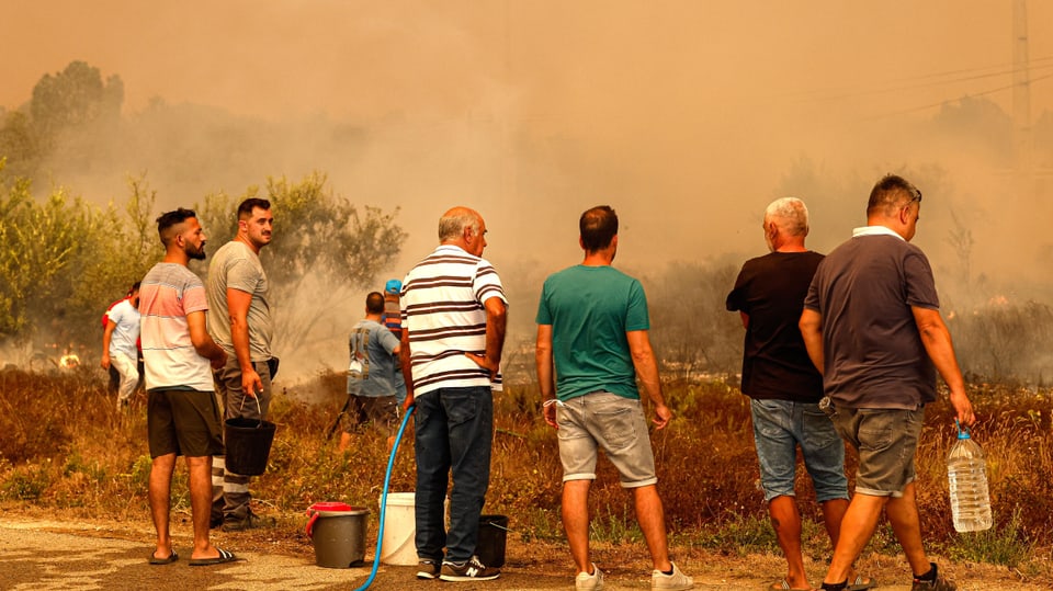 Mehrere Männer stehen, zum Teil mit Wasserbehältern in der Hand, vor einem Feld, aus dem Rauch steigt