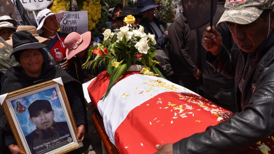 Viele Menschen stehen um einen Sarg, der mit einer rot-weissen Peru-Flagge umwickelt ist. Darauf sind Blumen.