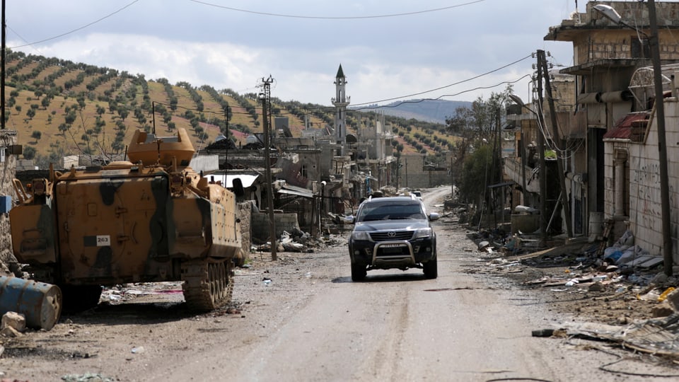 Panzer und Auto auf einer Strasse, daneben beschädigte Häuser, im Hintergrund Olivenplantagen.