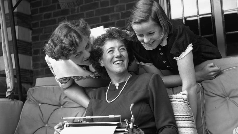Eine Frau auf dem Sofa, auf dem Schoss eine Schreibmaschine. Ihre beiden Töchter schauen ihr links und rechts über die Schulter.