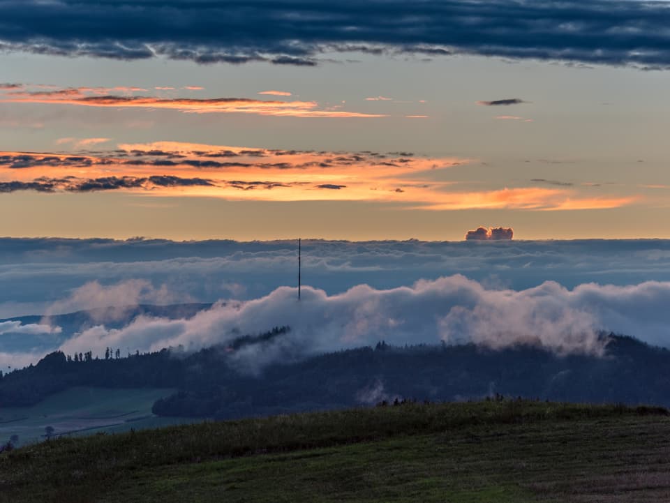 Eine tiefe Wolkenschicht umgibt den Sendeturm Bantiger. Hohe Wolkenfelder leuchten in der Abendsonne.