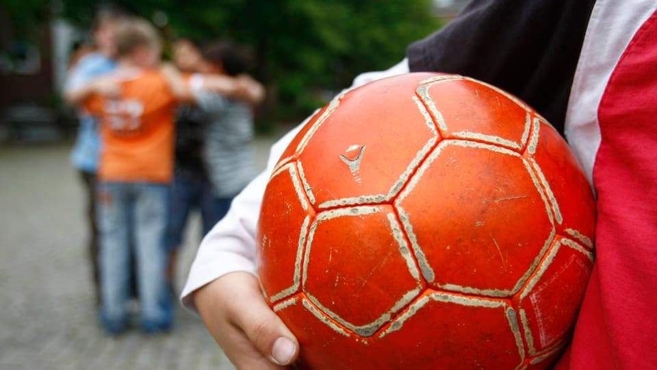 Ein Kind hält einen abgewetzten roten Fussball unter dem Arm. 