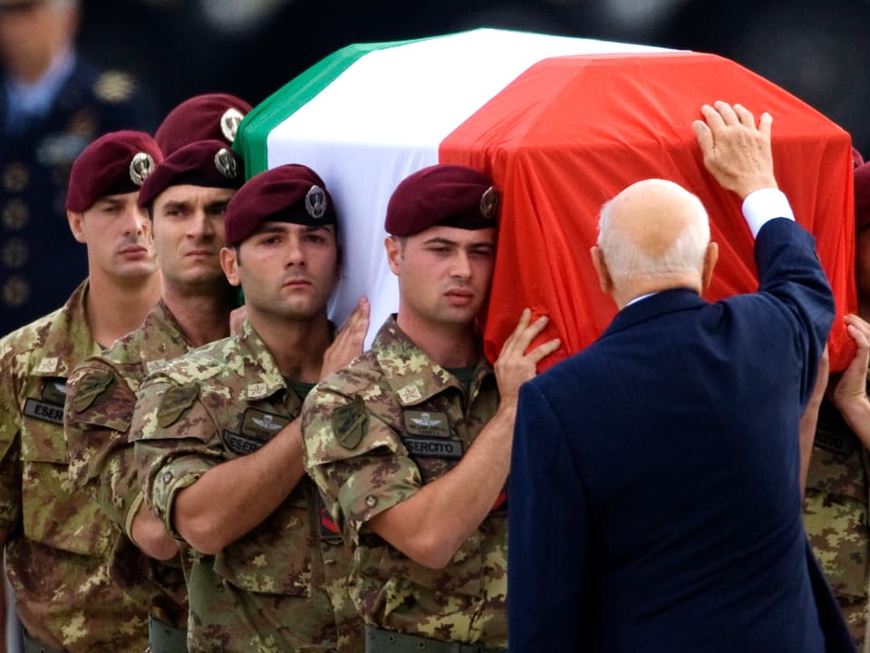 Napolitano hält eine Hand an einen Sarg, in dem ein getöteter Soldat liegt.