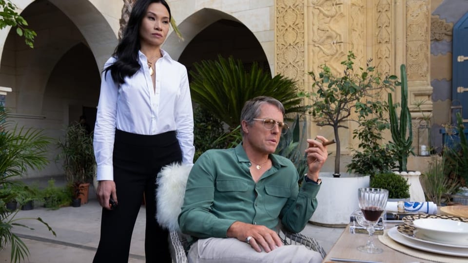 Auf Stuhl mit Fell sitzt ein Mann im grünen Hemd mit Zigarre und Sonnenbrille, hinter ihm steht junge asiatische Frau