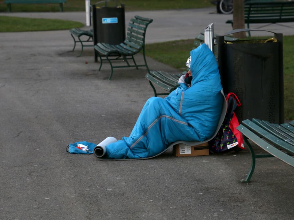 Ein Park in dem jemand auf einer Kartonkiste in einem blauen Schlafsack sitzt. Die Person ist am betteln und es ist kalt