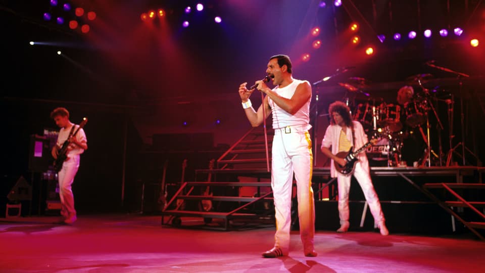 Freddie Mercury performt auf der Bühne in weisser Hose und Trägershirt.