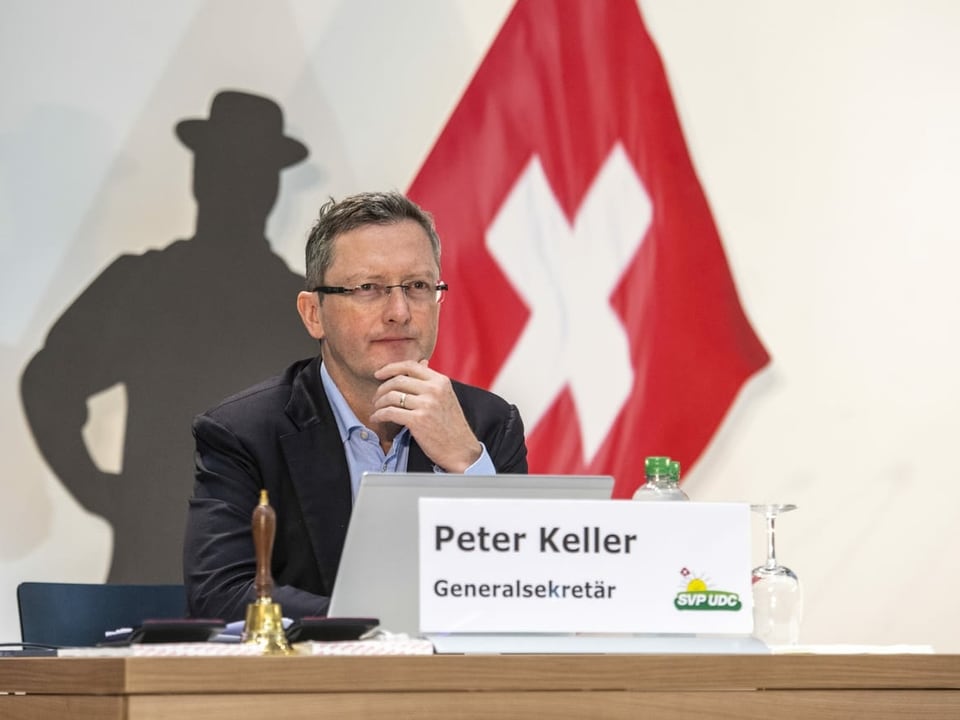 Ein Mann im Anzug vor einem Bild einer Schweizerfahne. Vor ihm steht ein Schild mit der Aufschrift "Peter Keller, Generalsekretär".